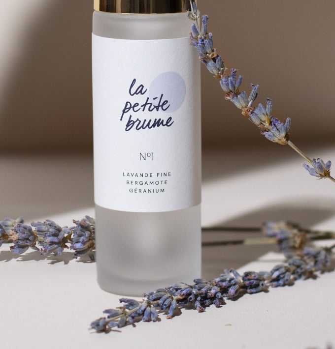 Un flacon de La Petite Brume avec du verre dépoli, un brin de lavande sur la droite et deux brins de lavande bleu devant le flacon. Sur un fond beige.